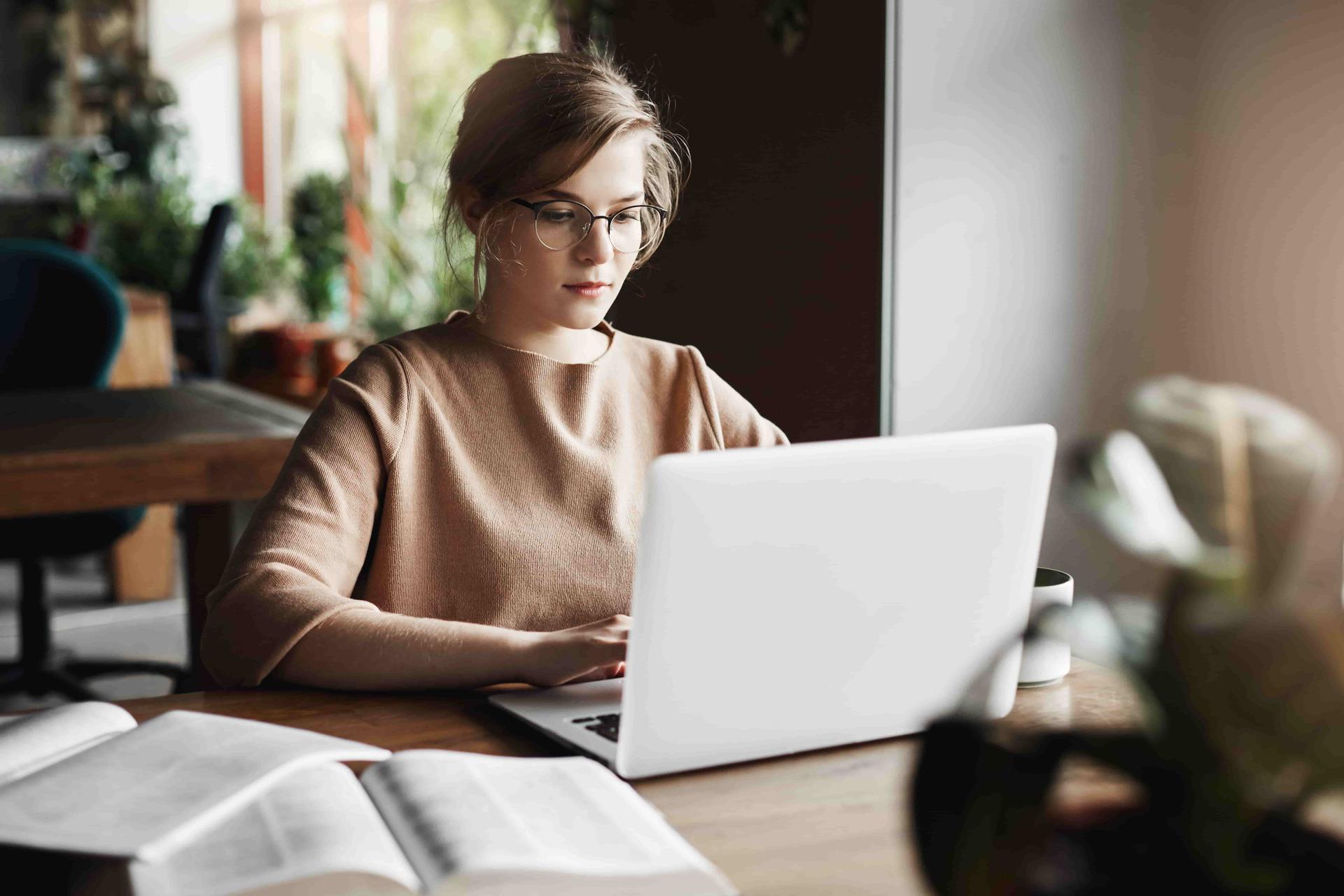 work-lifestyle-and-business-concept-good-looking-focused-european-female-in-trendy-glasses-sitting-in-cafe-near-laptop-working-on-notebook-surrounded-with-books-making-notes-min